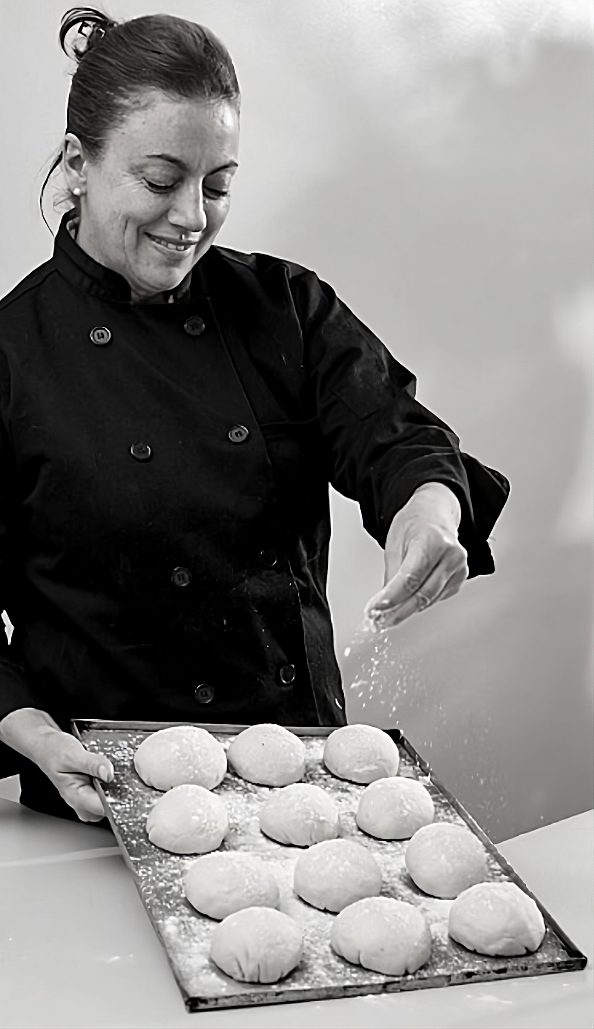 María Rosa, con chaqueta de chef negra, espolvoreando harina sobre una bandeja de bollos de masa